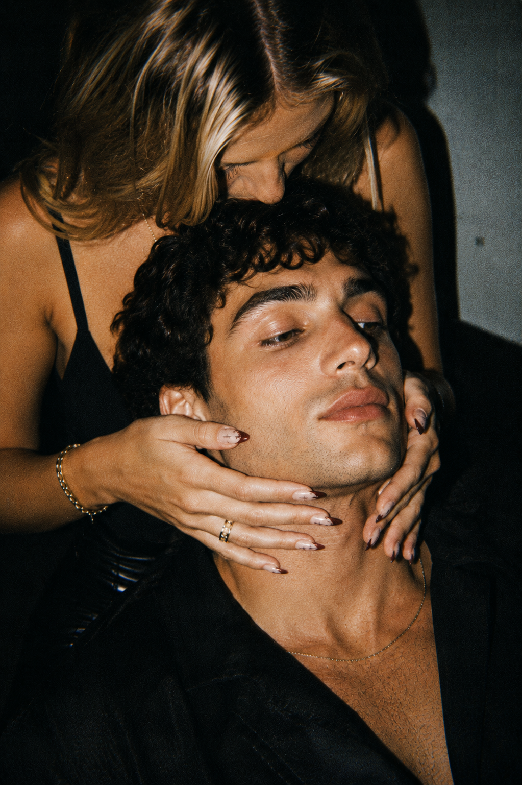 A close-up lifestyle shot of a man with curly dark hair resting his head while a woman with blonde hair gently holds his face. Both are wearing black clothing, set against a dark, moody background.
